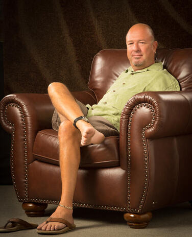 An image of Alvin Law sitting in a chair. He is wearing a t-shirt and shorts. His right leg is resting over his left knee, showing his bare foot. 